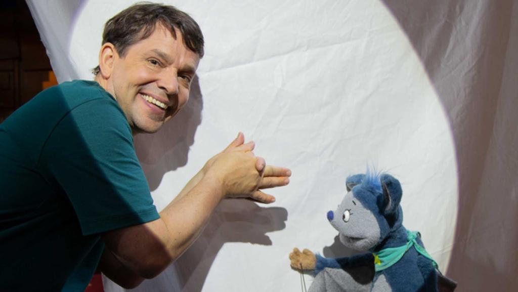 Juri hat ein weißes Stück Stoff aufgespannt und dahinter eine helle Lampe gestellt. Er zeigt Fidi ein paar Schattenspielfiguren, die er mit seinen Händen bildet.