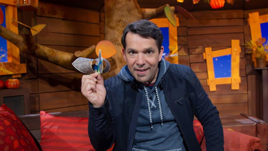 Moderator Juri sitzt im Baumhaus und hat einen gebastelten Flieger in der Hand.