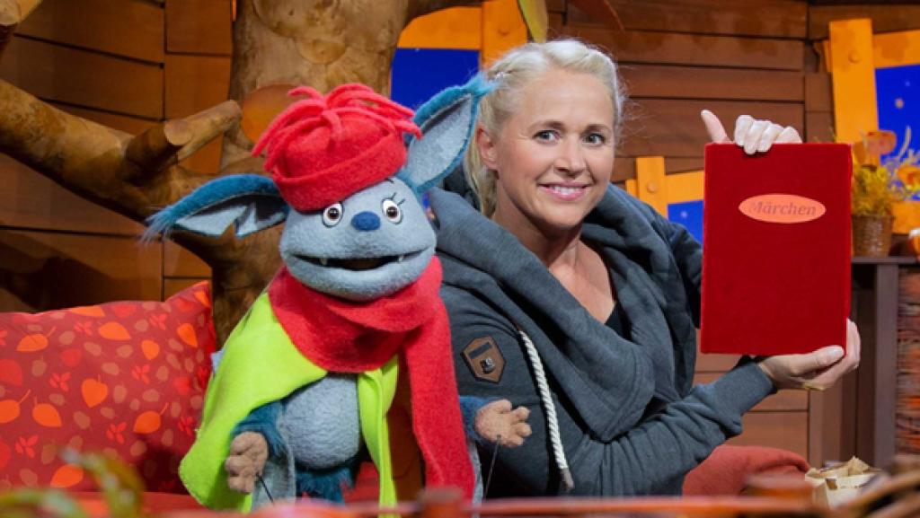 Fidi ist mit Mütze und Schal in rot bekleidet und Singa hält ein großes Märchenbuch in der Hand.