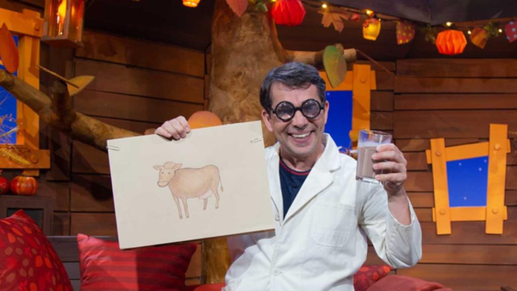 Professor Juri sitzt mit seinem weißen Kittel im Baumhaus auf der Bank. Er hält in der einen Hand ein Glas Kakaomilch und in der anderen ein Bild von einer gemalten Kuh hoch.