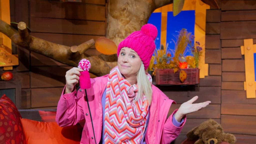 Singa trägt eine pinke Mütze und Regenjacke. In der rechten Hand hält sie eine kleine pinke Mütze.