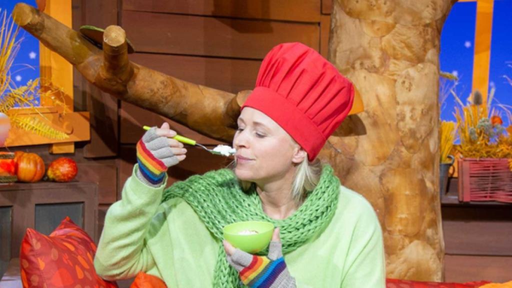 Singa trägt eine Kochschütze und eine Kochmütze. Sie riecht an einem Löffel Milchreis.