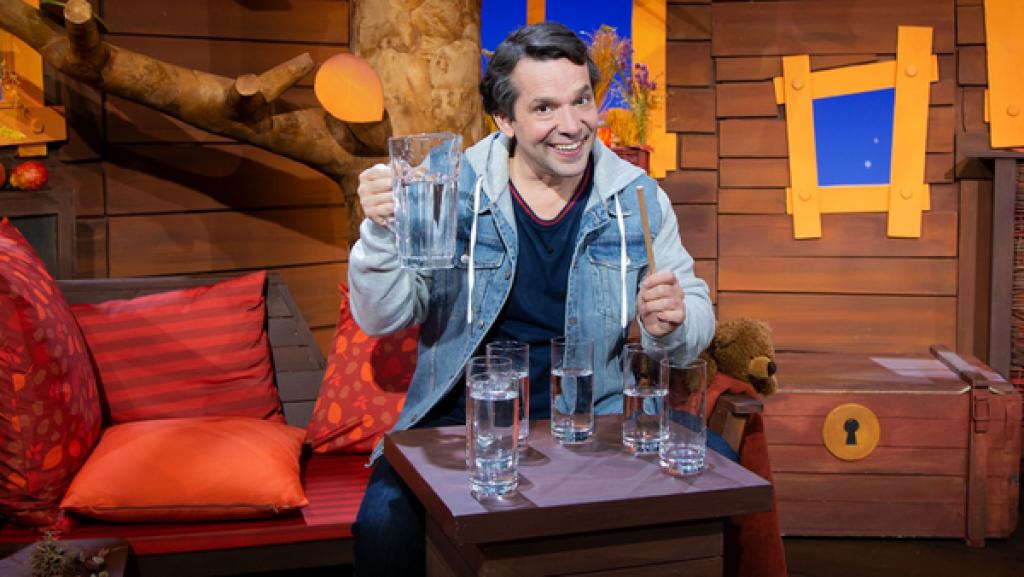 Juri hält eine Kanne mit Wasser und einen Holzstab. Vor ihm stehen sechs verschieden volle Gläser.