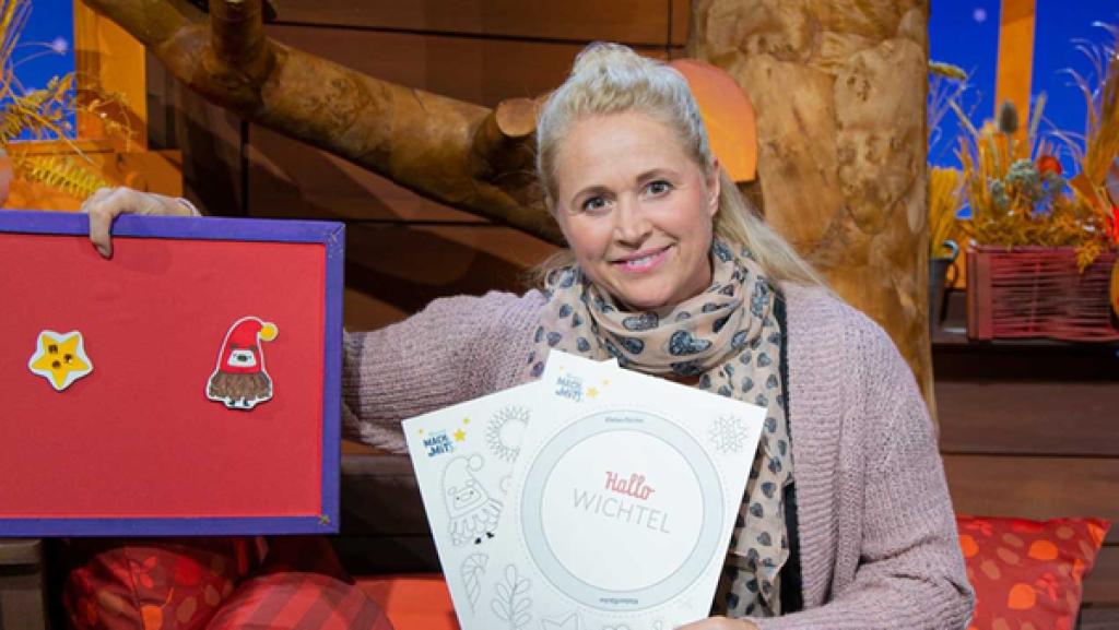 Singa zeigt den MACH MiT!-Bastelbogen. Auf dem Tisch liegen Bastelmaterialien, die sie braucht.