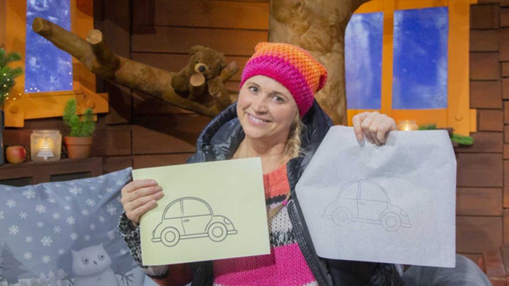 Singa hält ein Blatt Papier und ein Stück Backpapier in der Hand. Auf beiden Blättern ist ein Auto zu sehen.