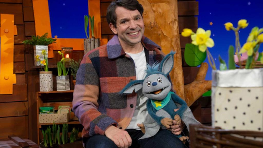 Rechte: KiKA Juri hat Fidi auf dem Schoß. Rechts im Bild steht ein Topf mit gelben Blumen.