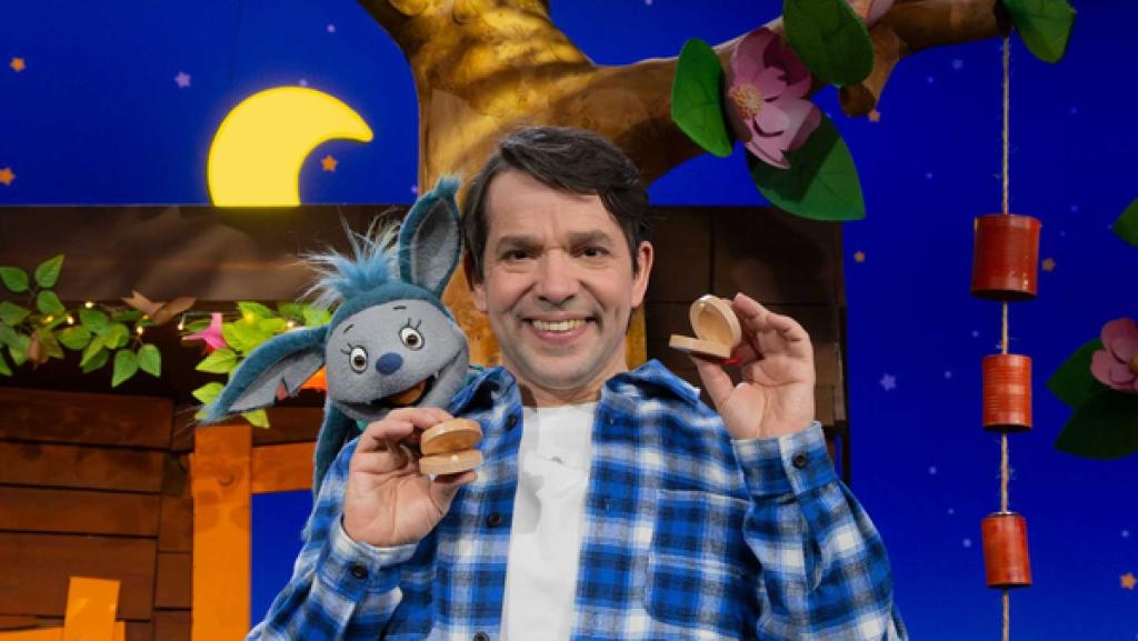 Rechte: KiKA Juri steht im Baumhaus. In jeder Hand hält er Klappern aus Holz. Fidi schaut über seiner linken Schulter hervor.