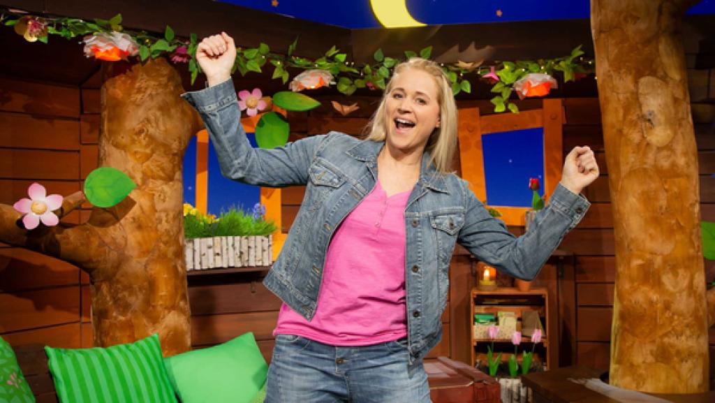 Rechte: KiKA Singa posiert mit Jeansjacke im Baumhaus. Sie lacht und hat die Arme fröhlich nach oben gestreckt.