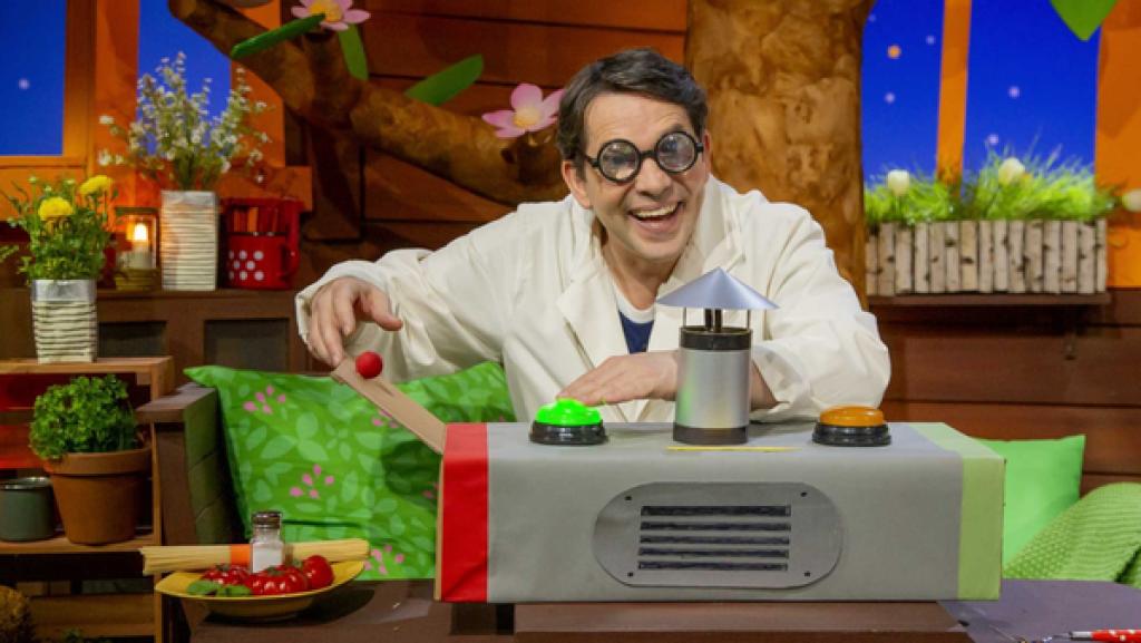 Rechte: KiKA Professor Juri sitzt hinter einem Automat und drückt auf eine leuchtende Lampe.