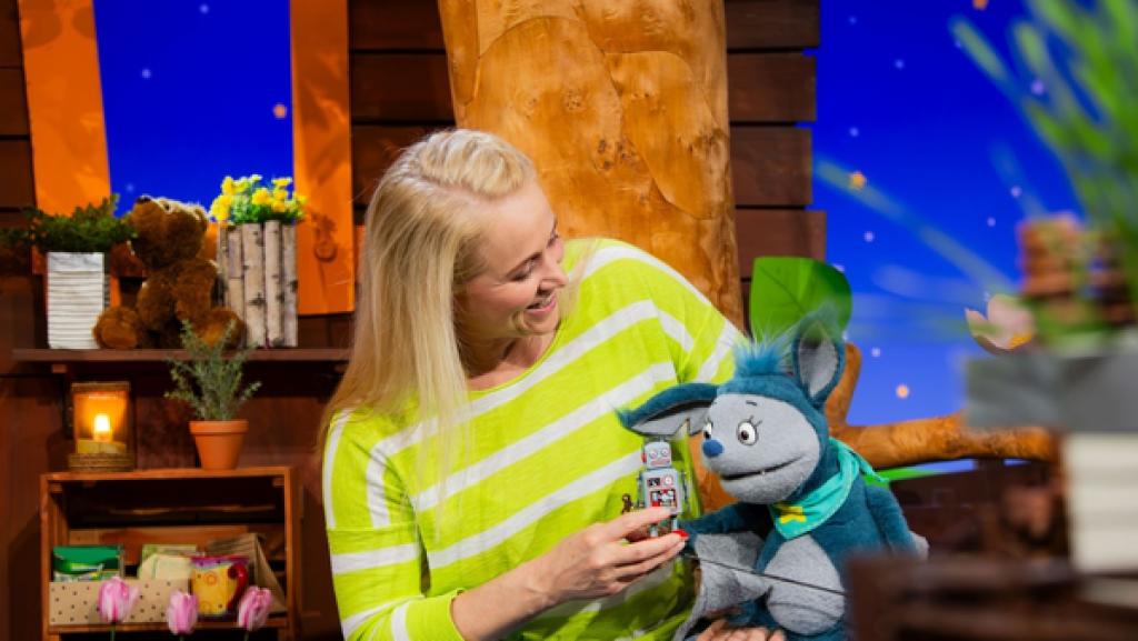 Rechte: KiKA Fidi sitzt auf dem Schoß von Singa, die einen kleinen Roboter in der Hand hält.