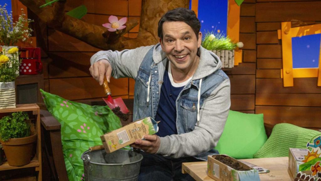 Rechte: KiKA Juri hält einen Milchkarton mit einem breiten Loch in der Hand. In diesen schaufelt er Erde hinein.