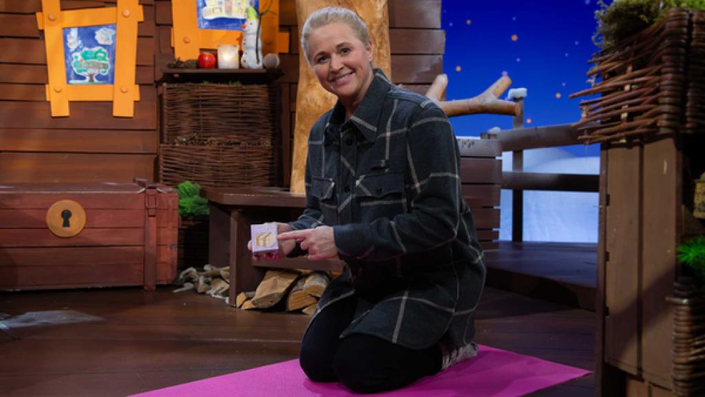 Singa kniet auf einer rosa farbenen Sportmatte auf dem Baumhausboden und zeigt auf den Ruhewürfel.