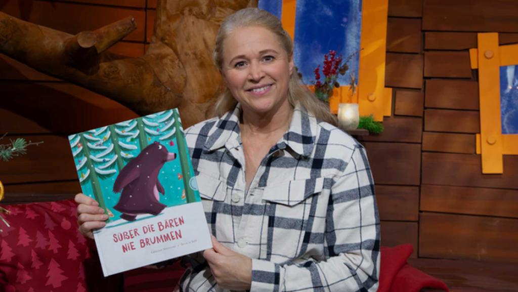 Singa sitzt auf dem Sofa und hält das Buch "Süßer die Bären nie brummen" hoch.