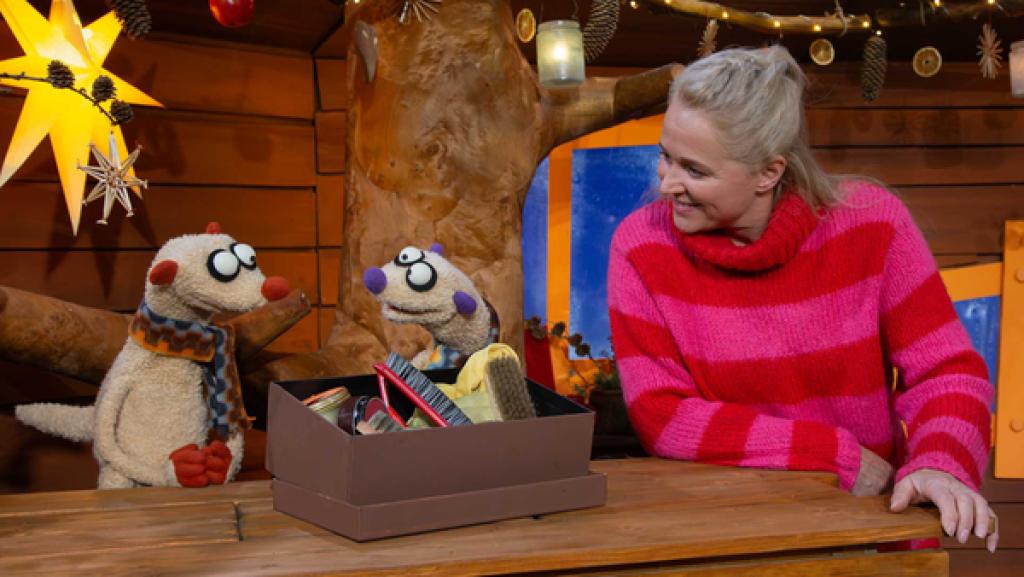 Singa schaut lachend zu den Erdmännchen Jan und Henry. Vor ihnen steht eine Box mit Schuhputzzeug.