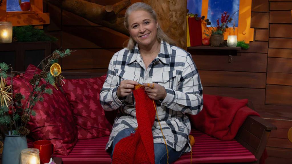 Singa sitzt auf dem Sofa und strickt mit roter Wolle einen Schal, der über ihrem Knie liegt.