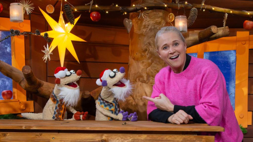 Singa zeigt lachend auf die Erdmännchen Jan und Henry. Sie tragen Weihnachtsmützen und weiße Bärte.