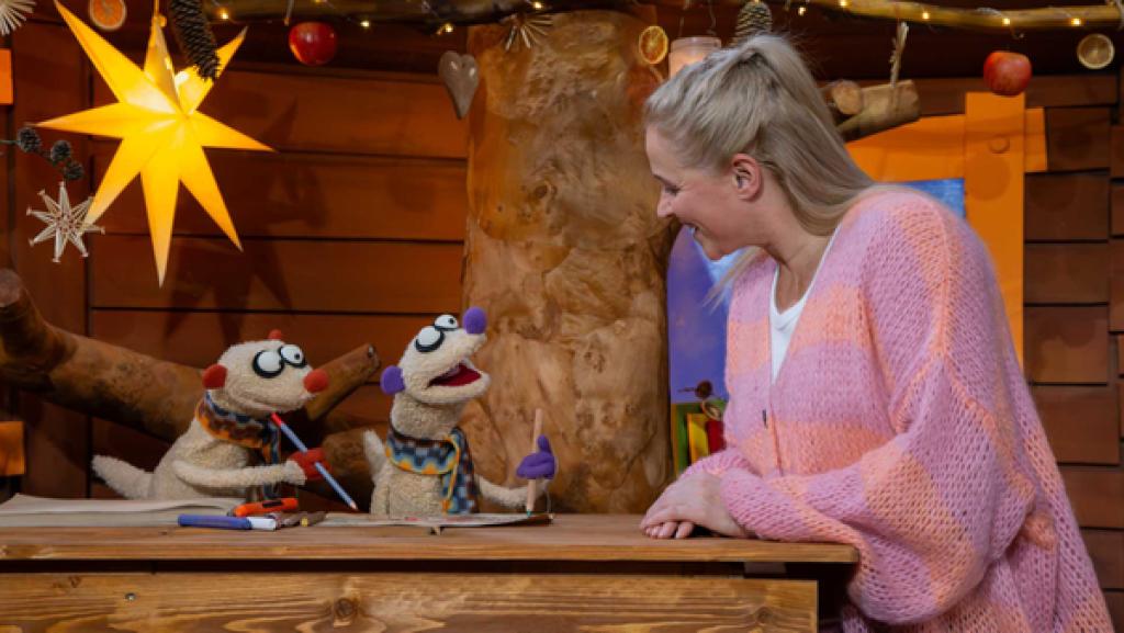 Singa steht bei den Erdmännchen Jan und Henry im Baumhaus. Die beiden halten Stifte und malen.