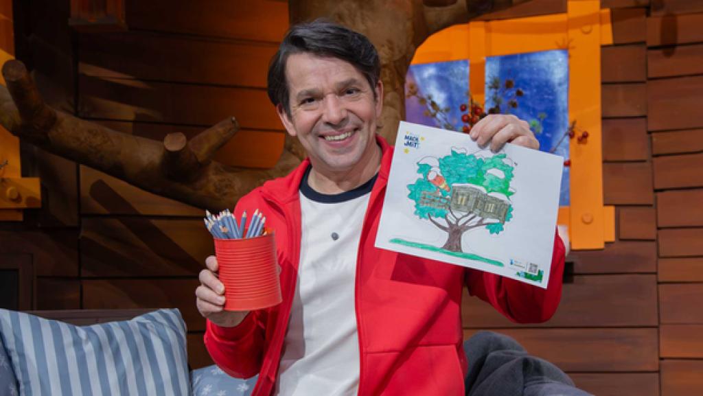 Juri hält lachend ein ausgemaltes MACHMiT!-Fensterbild und bunte Stifte in den Händen.