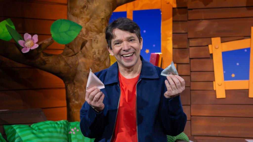 Juri sitzt im Baumhaus, lächelt und hält kleine Tüten mit Süßigkeiten in den Händen.