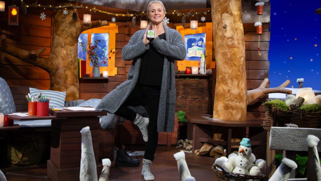 Singa steht im Baumhaus auf einem Bein. Das andere Bein winkelt sie nach außen gestreckt an. Der Ruhewürfel ist in ihrer Hand.