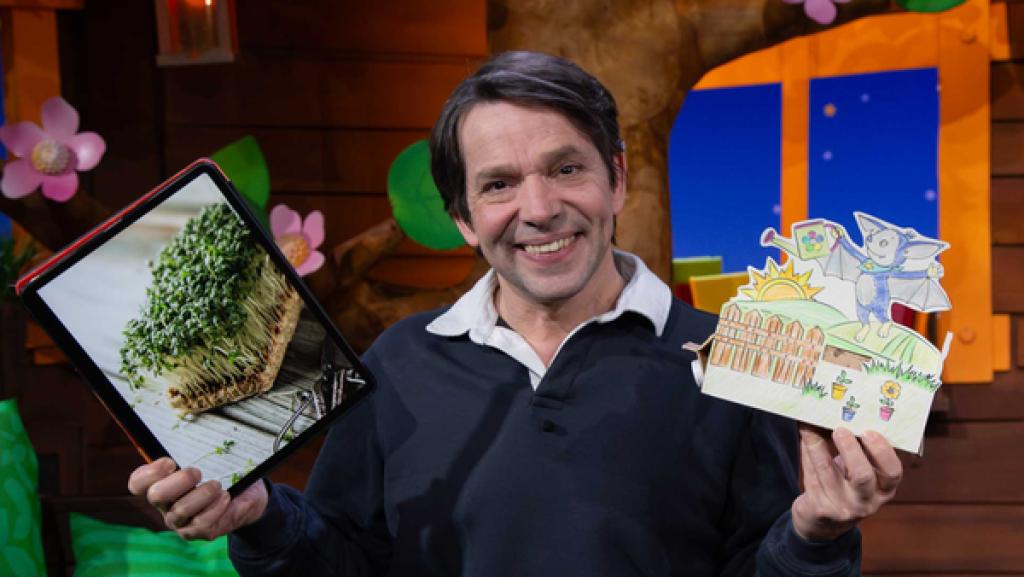 Juri sitzt im Baumhaus und hält in der linken Hand ein Kresse-Bild und rechts einen Fidi Garten.