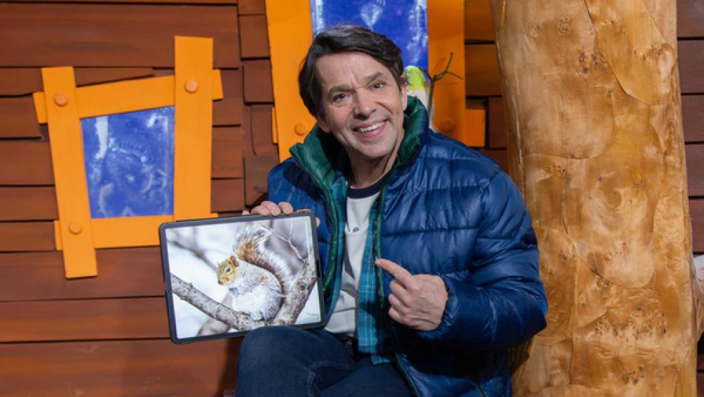 Juri sitzt lachend im Baumhaus und zeigt auf dem Tablet ein Bild von einem Eichhörnchen im Schnee.