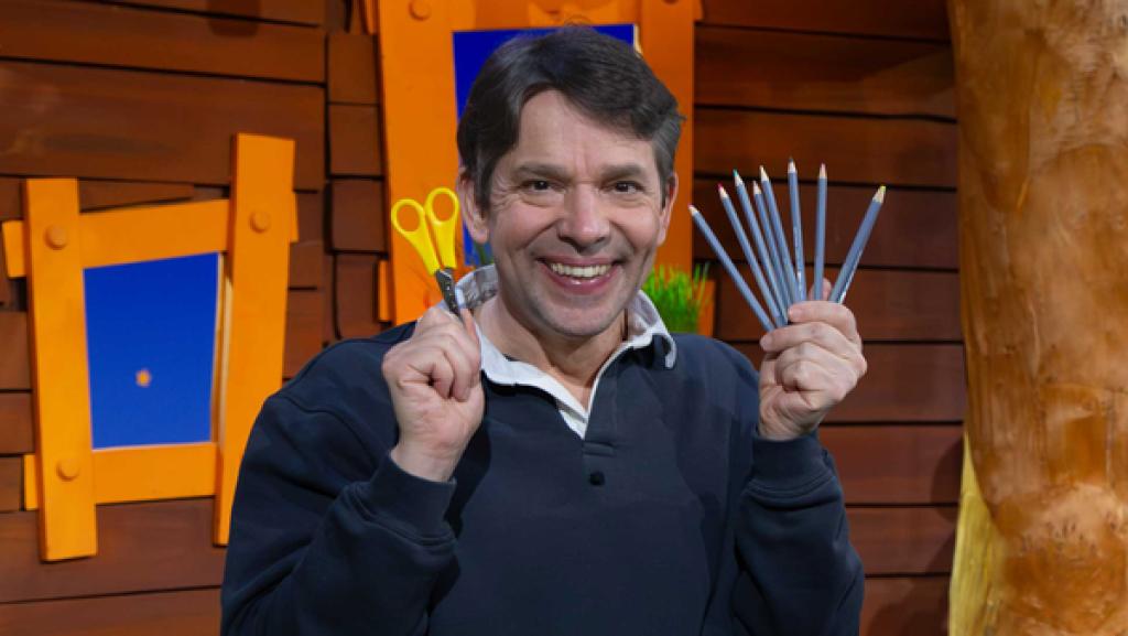 Juri steht im Baumhaus und hält in der einen Hand eine Schwere und in der anderen Hand bunte Stifte.