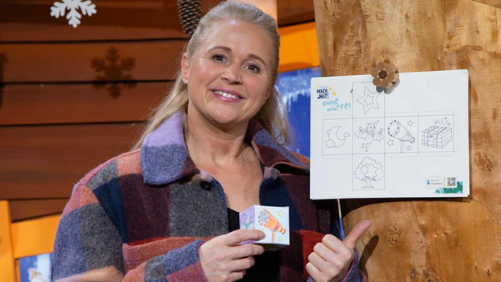 Singa steht neben dem Baumhausbaum, am Stamm hängt ein Bastelbogen mit Würfelmuster.