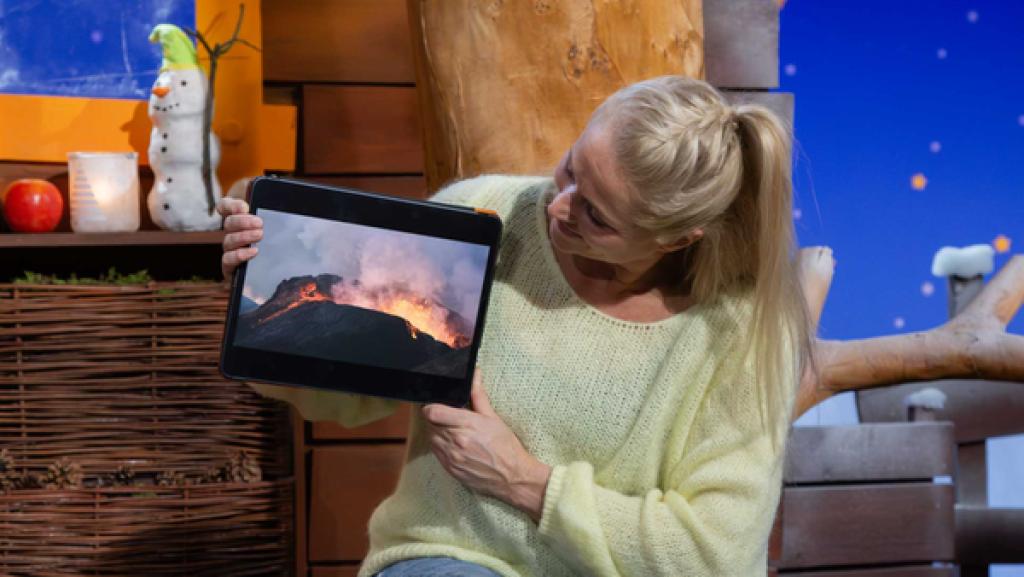 Singa sitzt im Baumhaus und zeigt auf dem Tablet ein Bild von einem Vulkan.