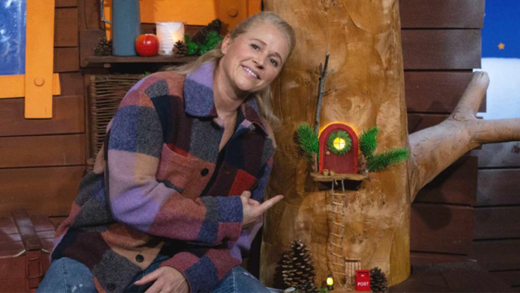 Singa sitzt am Baumstamm des Baumhausbaumes und zeigt auf das Wichteltürchen.