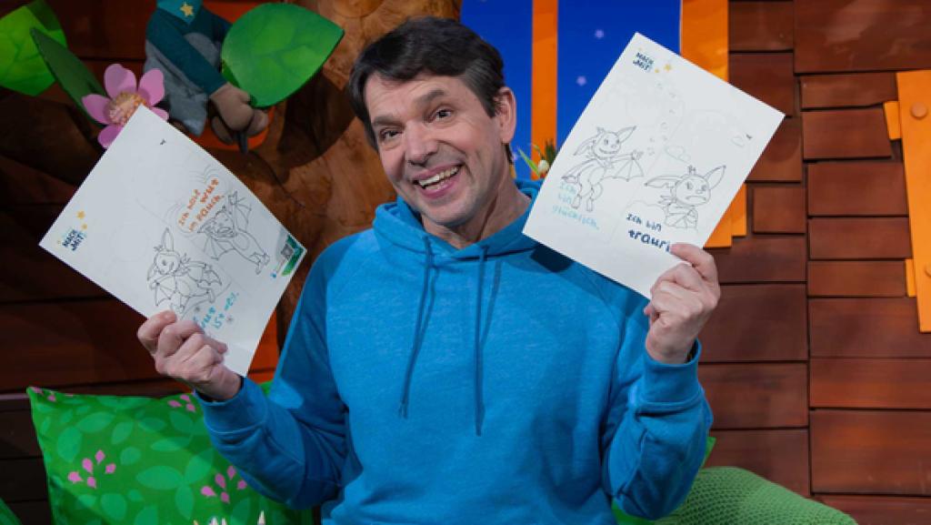 Juri steht im Baumhaus und hält in den beiden Händen die neuen Mach-Mit Bastelbögen für das Türschild.