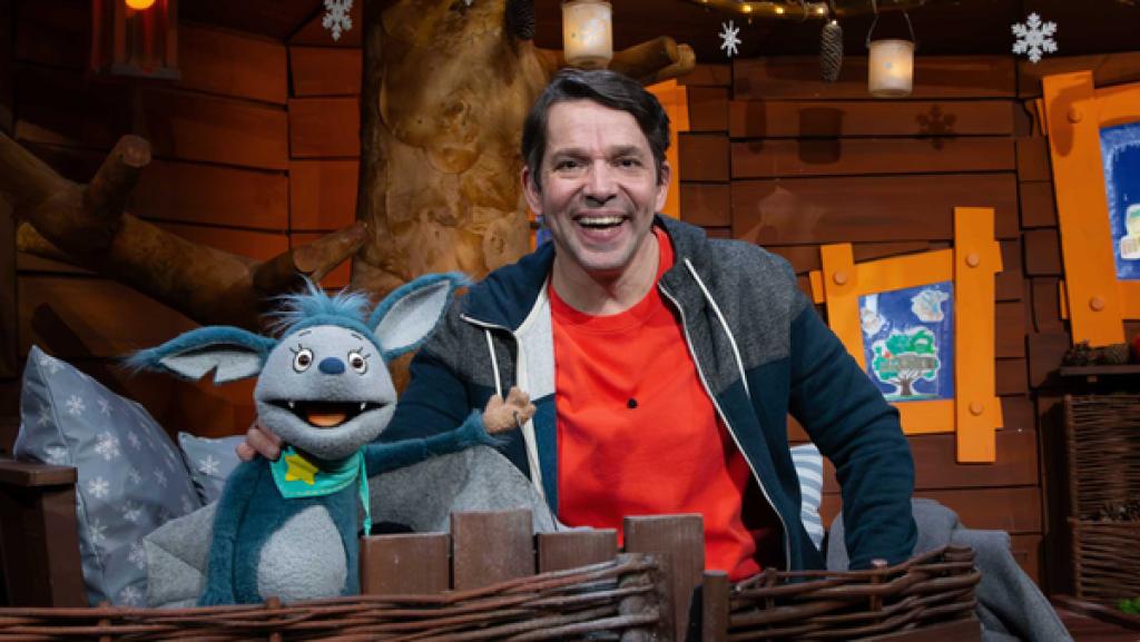 Juri und Fidi sitzen lächelnd auf dem Sofa im Baumhaus. Fidi winkt.