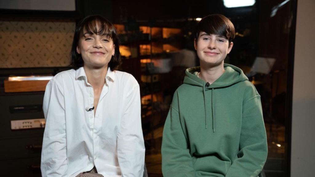 "Dein Song"-Kandidatin Leni (16) und Musikpatin Lotte sitzen nebeneinander in einem Tonstudio. Dabei blicken sie freundlich lächelnd in die Kamera.