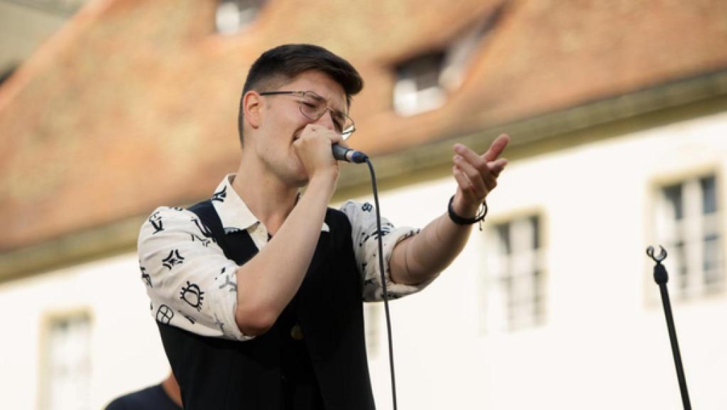 „Dein Song“-Kandidat Nikita (17) steht auf einer Freilichtbühne und singt leidenschaftlich in ein Mikrofon, das er in seiner Hand hält. Mit der anderen Hand macht er eine pathetische Geste nach vorne.