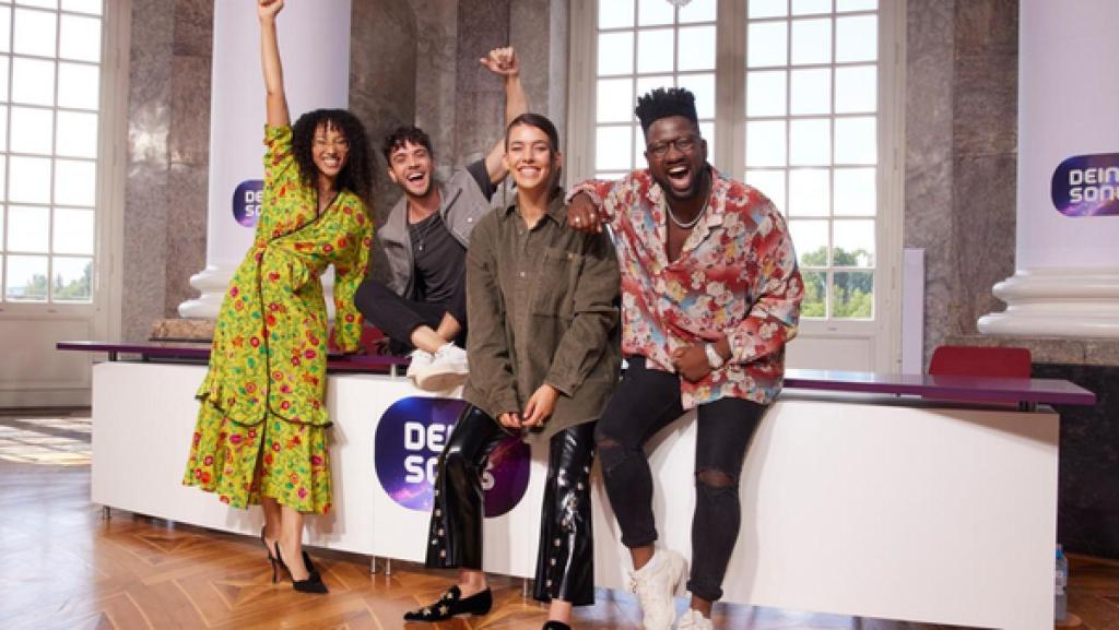 Die vier Jury-Mitglieder der 15. Staffel Dein Song stehen an ein Pult mit Dein Song Logo angelehnt und blicken fröhlich und lachend in die Kamera.