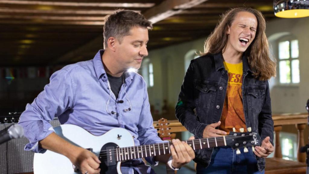 Kandidat Leon (14) steht auf der „Dein Song“-Probebühne und singt leidenschaftlich. Links neben ihm sitzt der Gitarrist der „Dein Song“-Band und begleitet Leon an der Gitarre.
