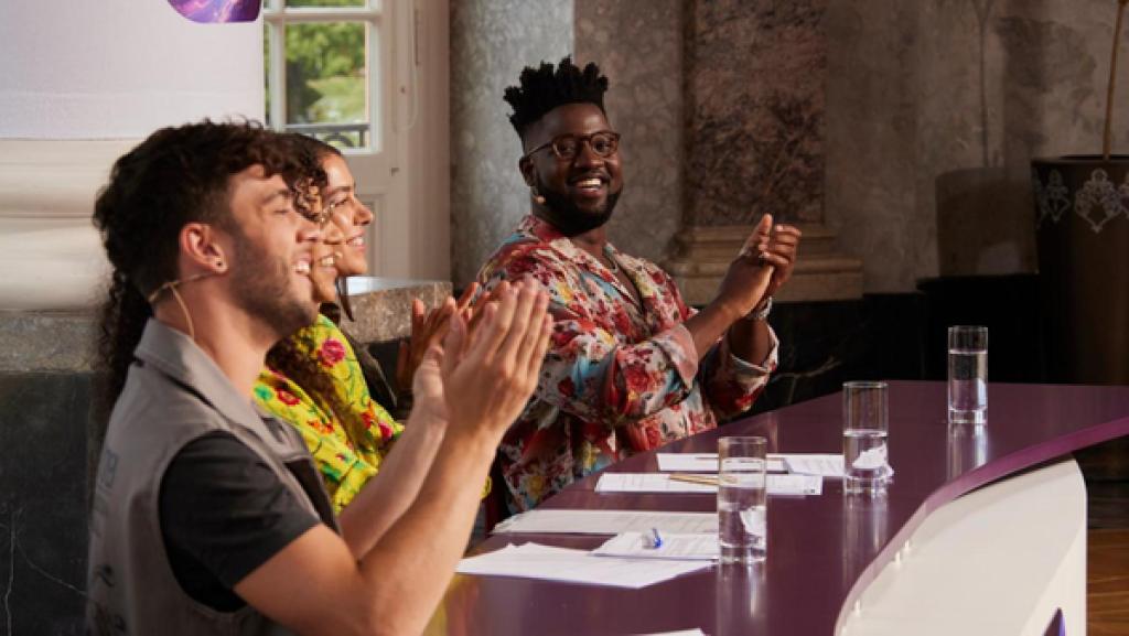 Die Jury-Mitglieder Luca Hänni, Suena, Alina Süggeler und Kelvin Jones sitzen hinter ihrem Jury-Pult, auf dem mehrere weiße Blätter verteilt liegen. Dabei lächeln und applaudieren sie begeistert.