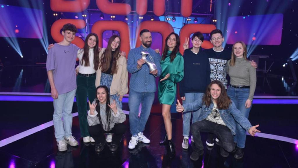 Die Finalisten und Finalistinnen sowie das Moderations-Duo der 15. "Dein Song"-Staffel stehen auf der "Dein Song"-Bühne und lächeln in die Kamera.