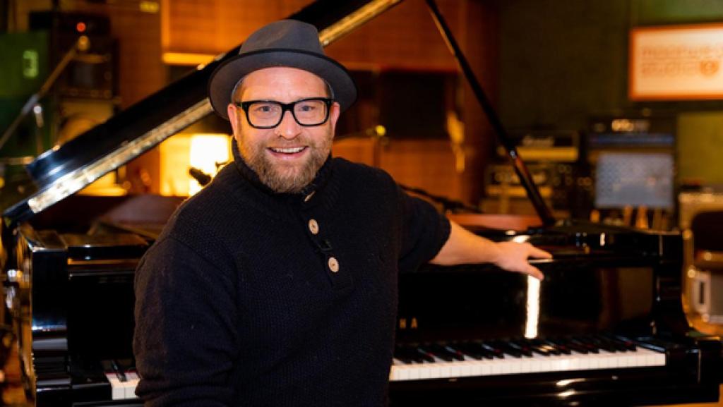 "Dein Song"-Musikpate Gregor Meyle sitzt im Tonstudio vor einem Klavier. Er lächelt freundlich in die Kamera. Seine linke Hand legt er dabei auf dem Klavier ab.