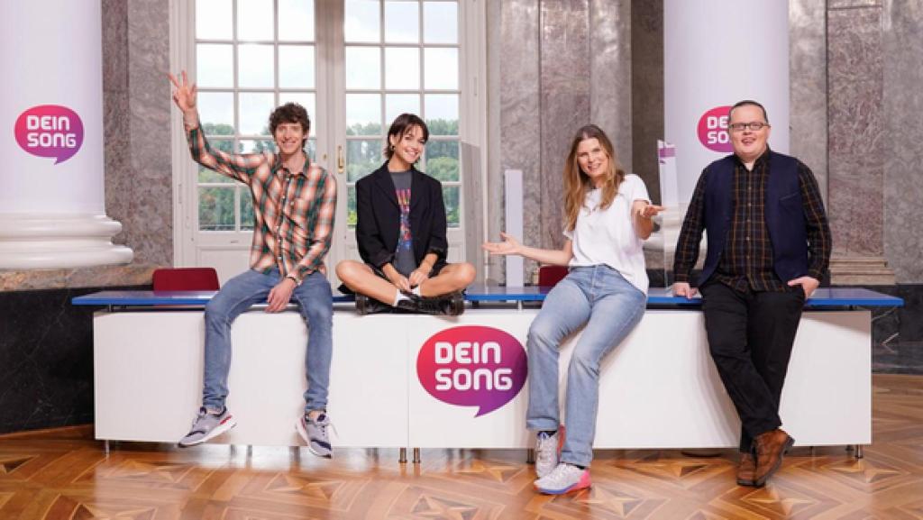 Dein Song 2022 Staffel 14: Die "Dein Song" Jurymitglieder v.l.n.r. Ole Specht, Lotte, Eva Briegel, Angelo Kelly sitzen und stehen vorm Casting-Pult und lachen in die Kamera. In der Mitte am Casting-Pult ist das "Dein Song"-Logo in weißer Schrift in Form von einer lila/roten Sprechblase zu sehen.