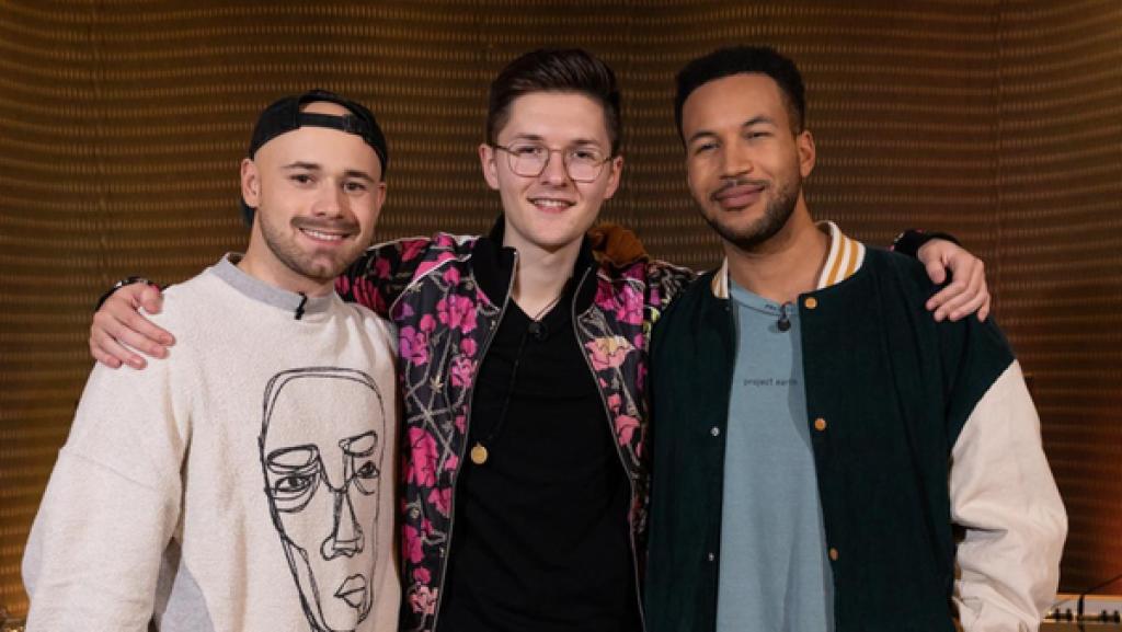 "Dein Song"-Kandidat Nikita (17) und die Musikpaten Josh Tapen und Janik Riegert stehen im Tonstudio vor einem Mischpult. Nikita steht in der Mitte und hat seine Arme um die beiden Musikpaten gelegt. Alle drei blicken freundlich in die Kamera.