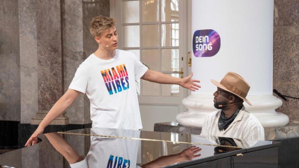 Juror Kelvin Jones sitzt am Klavier und blickt freundlich zu Kandidat Marc. Marc steht neben dem Klavier und breitet seine Arme aus, während er gefühlvoll singt.