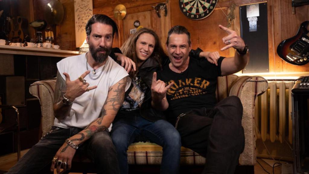 "Dein Song"-Kandidat Leon (14) und Sascha Vollmer und Alec Völkel von "The Boss Hoss" sitzen in einem Saloon auf dem Sofa. Sie lachen dabei motiviert in die Kamera. Mit ihren Händen formen sie das Heavy-Metal-Zeichen.