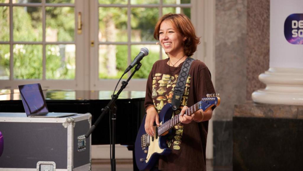 Kandidatin Estrela (18) steht lächelnd auf der "Dein Song"-Bühne und hält eine Gitarre in den Händen. Vor ihr steht ein Mikrofon. Links im Hintergrund steht ein schwarzer Flügel. Durch das weiße sprossen Fenster hinter dem Flügel, kann man den Schlosspark sehen.