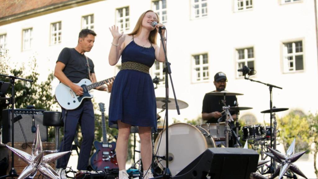 "Dein Song"-Kandidatin Hanna (16) steht auf einer Freilichtbühne und singt gefühlvoll in ein Mikrofon. Im Hintergrund ist auch die "Dein Song"-Band zu erkennen. Links und rechts neben Hanna sind dekorative, silberne Sterne zu sehen.