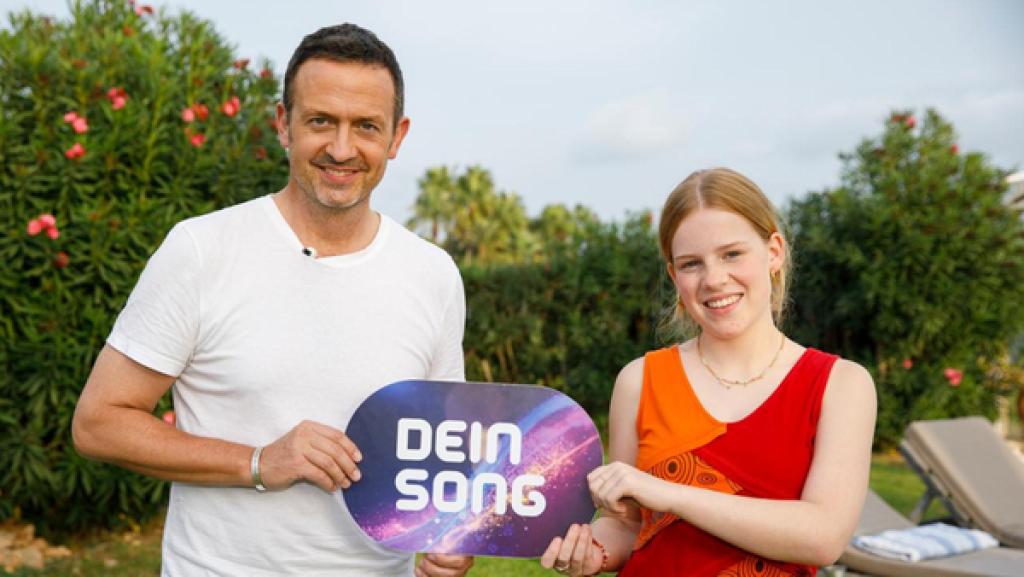 Till Brönner und Frida stehen nebeneinander und halten ein Dein Song-Schild zwischen sich. Sie lächeln.