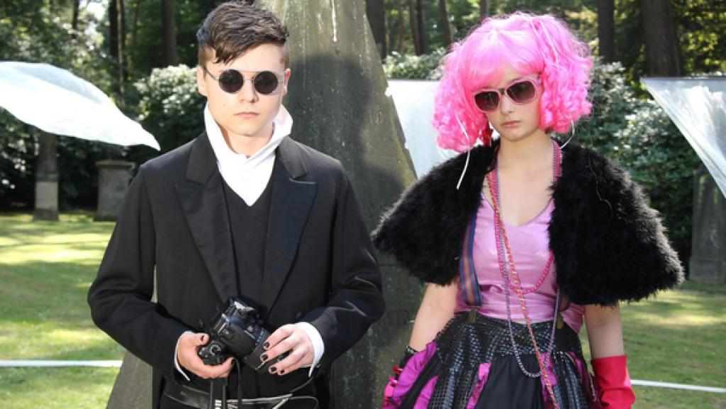 Ein Junge (Tom Böttcher) und ein Mädchen (Johanna Werner) mit Sonnenbrillen und auffälliger Kleidung stehen unter Bäumen.