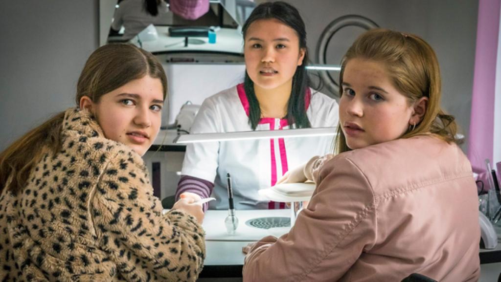 Lou, Mai (My Dam) und Pippa müssen vorsichtig sein; sie stehen unter Beobachtung. | Rechte: NDR/Letterbox, Boris Laewen Lou und Pippa sitzen an einem Tisch im Nagelstudio, auf der anderen Seite des Tisches sitzt Mai.