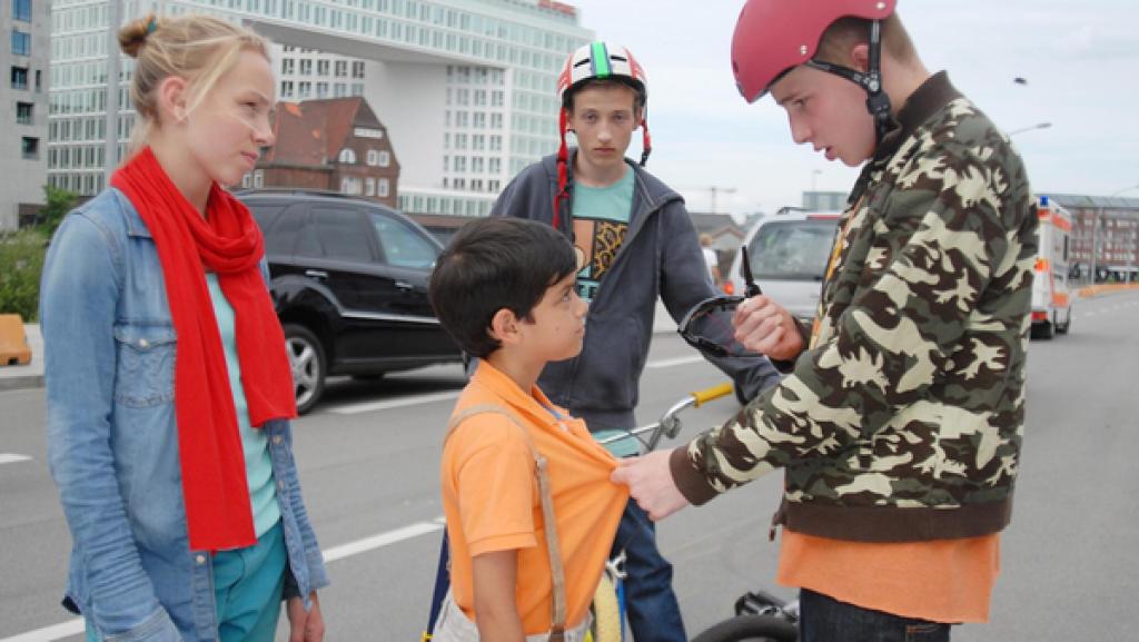 Jessi (Martha Fries) schaut zu einem Jungen mit BMX-Helm (David Riske), der eine Sonnenbrille in der Hand hält und Luis (Emilio Sanmarino, Mitte) am T-Shirt packt. Ein weiter Junge mit Helm schaut zu (Benjamin Falk, hinten)