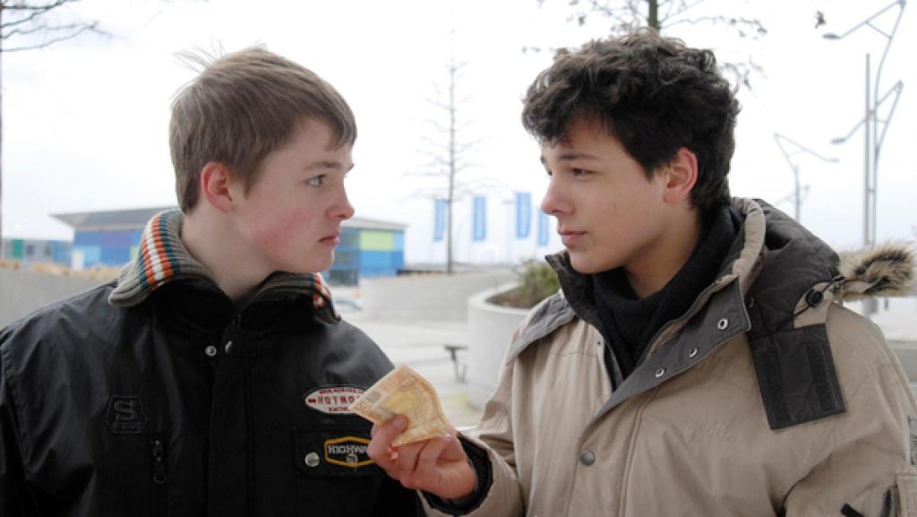 Ein Jugendlicher (Lukas Kieback, rechts) hält einem anderen (Maximilian Werner) einen Geldschein hin. Sie sehen sich an.